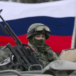 A pro-Russian man holds a Russian flag behind an armed servicemen on top of a Russian army vehicle outside a Ukrainian border guard post in the Crimean town of Balaclava